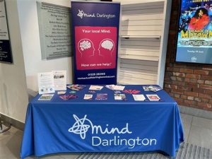 Photograph of a promotional table setup for Mind Darlington, a mental health support organization, featuring a blue tablecloth with the organization's logo and name. The table displays informational brochures, flyers, and a standing banner with the message "Your local Mind. How can we help?"
