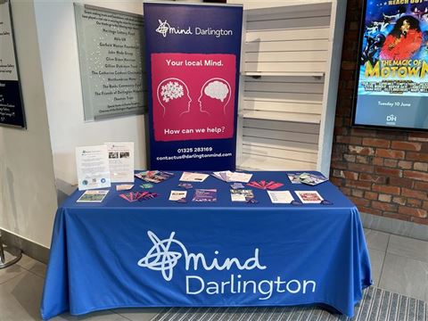 Photograph of a promotional table setup for Mind Darlington, a mental health support organization, featuring a blue tablecloth with the organization's logo and name. The table displays informational brochures, flyers, and a standing banner with the message "Your local Mind. How can we help?"