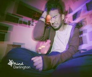 Photograph of a person sitting cross-legged indoors with a hand on their head, surrounded by a purple and pink light effect creating a blurred, dreamy atmosphere.