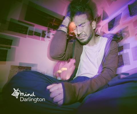 Photograph of a person sitting cross-legged indoors with a hand on their head, surrounded by a purple and pink light effect creating a blurred, dreamy atmosphere.
