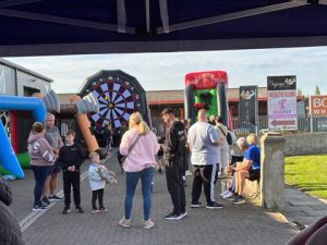 Photograph of an outdoor event with people gathered around inflatable games, including a large dartboard and a boxing glove target.