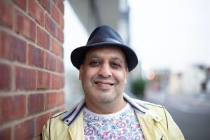 Photograph of a person standing outdoors next to a red brick wall wearing a black hat, yellow jacket, and a white shirt with a colorful paisley pattern.