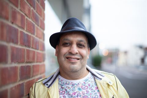 Photograph of a person standing outdoors next to a red brick wall wearing a black hat, yellow jacket, and a white shirt with a colorful paisley pattern.