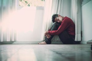 Photograph of a person sitting on a wooden floor in a corner, hugging their knees while wearing a red sweater and dark trousers.