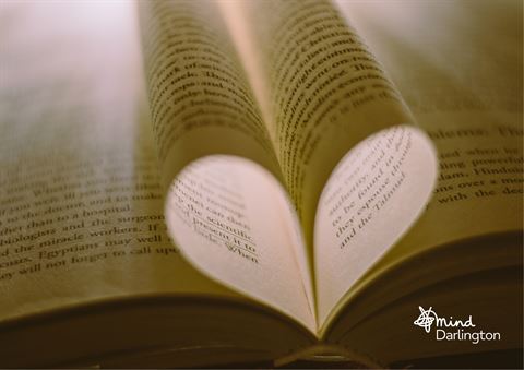 Photograph showing pages of an open book folded inward to form a heart shape at the center. Warm lighting highlights text on yellowed pages, creating a cozy and romantic atmosphere.