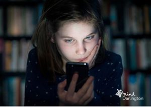 Photograph of a child holding a smartphone close to their face, with a background of blurred bookshelves.