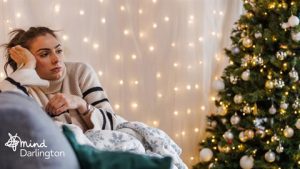 Photograph of a person sitting on a couch wrapped in a blanket with a decorated Christmas tree and warm string lights in the background.