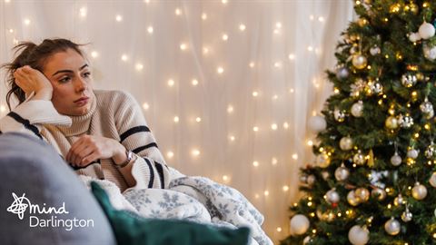Photograph of a person sitting on a couch wrapped in a blanket with a decorated Christmas tree and warm string lights in the background.