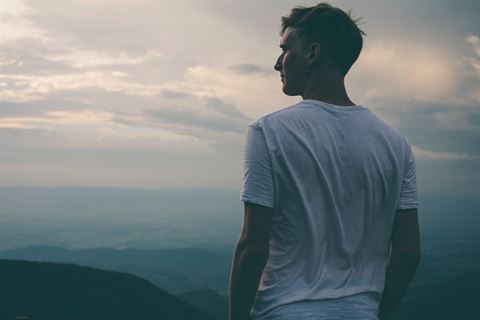 Photograph of a person standing and looking out over a vast mountainous landscape during a cloudy sunset.