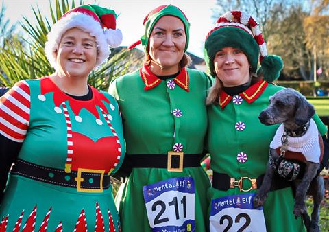 Photograph of three people dressed in festive elf costumes with green dresses, red and white striped sleeves, and black belts, standing outdoors.