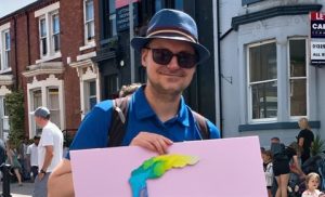 Photograph of a person wearing a blue shirt and hat holding a pink poster with a colorful abstract design in an outdoor urban setting.