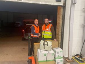 Photograph of two workers wearing orange and yellow safety vests standing next to a pallet jack loaded with cardboard boxes and a white insulated bag inside a warehouse loading dock.