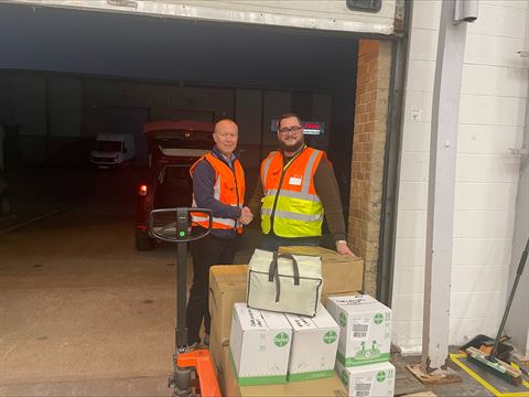 Photograph of two workers wearing orange and yellow safety vests standing next to a pallet jack loaded with cardboard boxes and a white insulated bag inside a warehouse loading dock.
