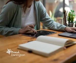 Photograph of a person using a tablet and laptop at a wooden desk, suggesting a work or study environment.