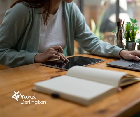 Photograph of a person using a tablet and laptop at a wooden desk, suggesting a work or study environment.