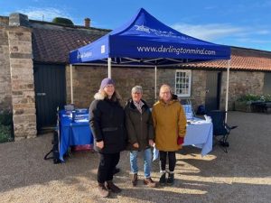Photograph showing three people standing in front of a blue canopy tent with "www.darlingtonmind.com"