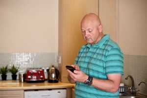 Photograph of a man standing in a kitchen holding a smartphone, wearing a teal and black striped polo shirt