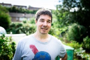 Photograph of a person standing outdoors in a garden or backyard setting with greenery and buildings in the background.