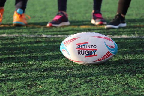 Photograph of a rugby ball on green grass. The ball features red and blue patterns and the text "GET INTO RUGBY,"