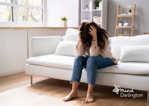 Photograph of a woman sitting barefoot on a white couch in a bright living room, holding her head in her hands, conveying stress or distress.
