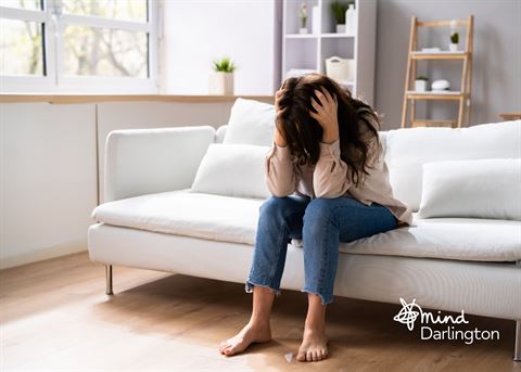 Photograph of a woman sitting barefoot on a white couch in a bright living room, holding her head in her hands, conveying stress or distress.