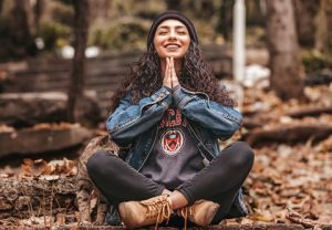 Photograph of a person sitting cross-legged on a log in an outdoor autumn setting, wearing a denim jacket, dark pants, tan boots, and headphones.