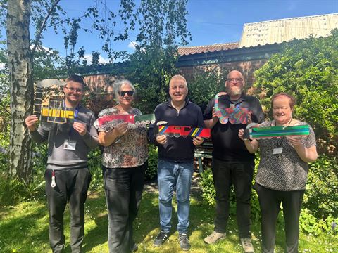 Photograph of five people standing outdoors in a garden holding colorful, uniquely designed cutouts shaped like vehicles or objects.