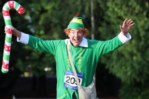 Photograph of a person dressed in a green elf costume with a striped candy cane prop, participating in an outdoor event. The costume includes a green hat with red accents and a race bib