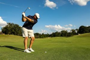 Photograph of a man preparing to swing a golf club on a green golf course under a partly cloudy blue sky. The man wears a white cap, navy shirt, and white shorts, with trees and a clear landscape in the background