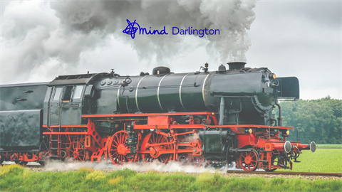 Photograph of a vintage black and red steam locomotive emitting thick gray smoke while traveling on railway tracks through a green field. The scene includes a cloudy sky and a "mind Darlington" logo