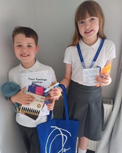 Photograph of two children standing side by side, each holding various school supplies and educational materials. The boy holds coloring pens, a stuffed toy, and a booklet titled "Your feelings matter," while the girl carries a blue tote bag, a laminated card on a lanyard, and a small orange object.