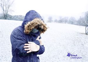 Photograph of a person outdoors in snowy landscape wearing a dark blue winter coat with fur-lined hood and white gloves, hugging themselves for warmth.