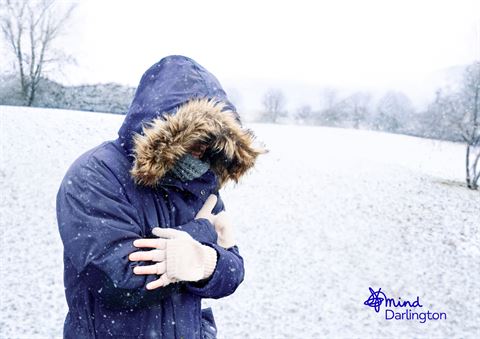 Photograph of a person outdoors in snowy landscape wearing a dark blue winter coat with fur-lined hood and white gloves, hugging themselves for warmth.