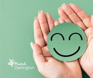 Photograph showing two hands gently holding a green circular cutout with a simple black smiling face on a matching green background. The image includes a small white logo and text "mind Darlington," suggesting a focus on mental health and well-being.