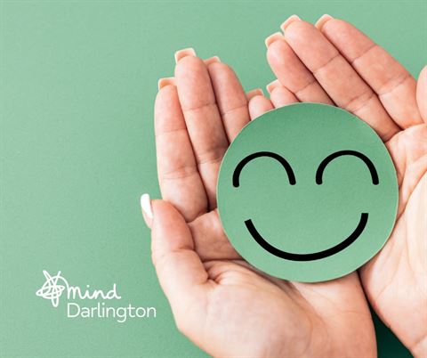 Photograph showing two hands gently holding a green circular cutout with a simple black smiling face on a matching green background. The image includes a small white logo and text "mind Darlington," suggesting a focus on mental health and well-being.