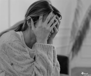 Photograph of a person sitting indoors wearing a thick knitted sweater, holding their head with both hands in a gesture suggesting stress or contemplation.