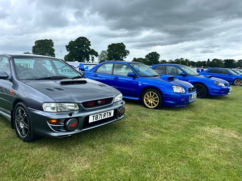 Photograph of a lineup of four Subaru cars parked on grass under a cloudy sky
